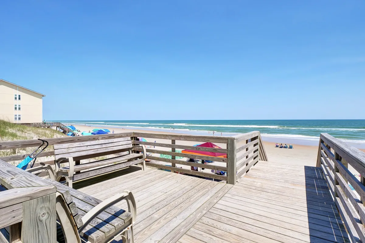 An inviting wooden deck overlooking the beach, with umbrellas and lounge chairs set up for relaxation. The ocean stretches beyond, offering a tranquil backdrop for morning coffee or sunset gatherings.