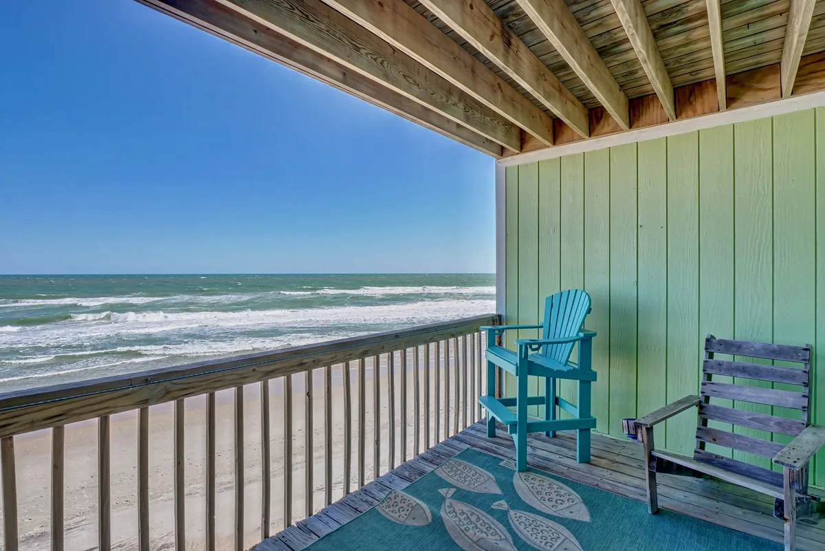 A cozy deck scene with a blue Adirondack chair and a wooden lounge chair set atop a decorative rug, all facing sweeping ocean views. The atmosphere is peaceful, inviting guests to relax, read, or socialize while soaking up the sights and sounds of the sea.