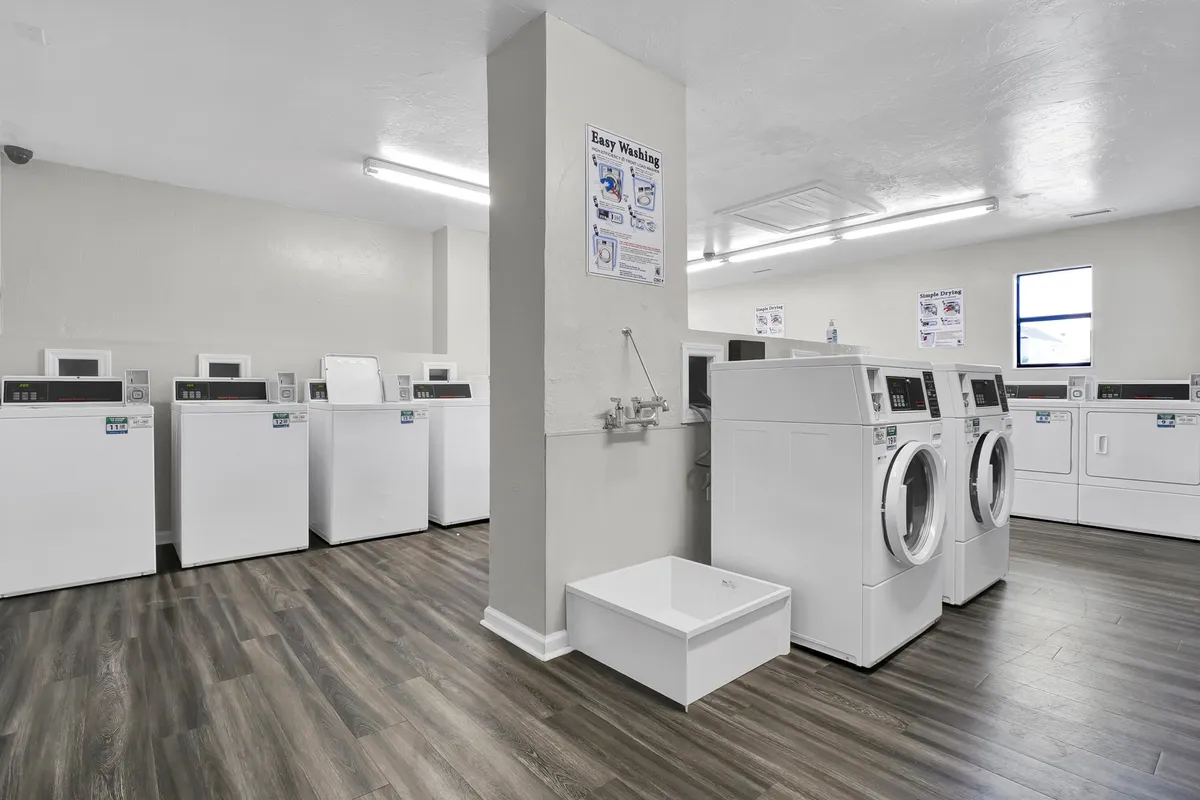 A clean and well-lit laundry facility equipped with multiple washing machines and dryers, signage on the wall, and space for guests to attend to their laundry needs. The mood is practical and welcoming.