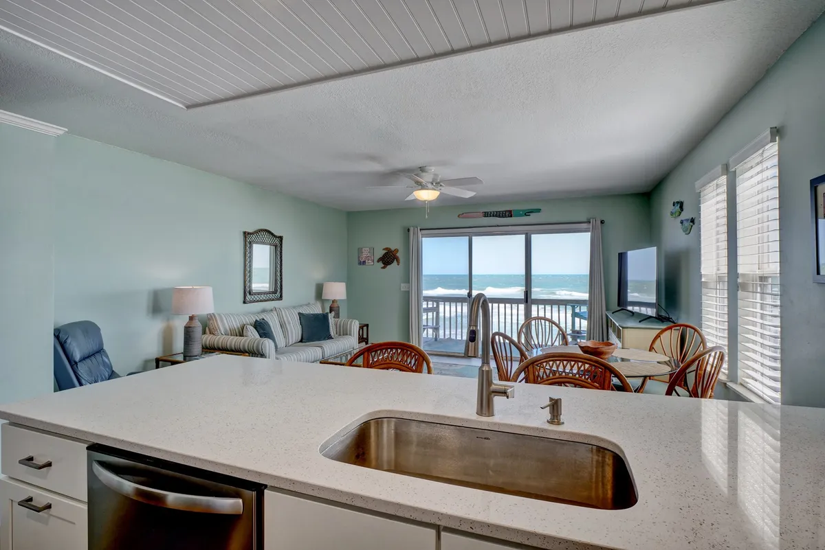 A welcoming living and dining area with large windows offering an ocean view. The space features a comfortable seating area, a dining table for four, and a television. Natural light floods the room, enhancing the cozy and communal atmosphere ideal for relaxing or sharing a meal.