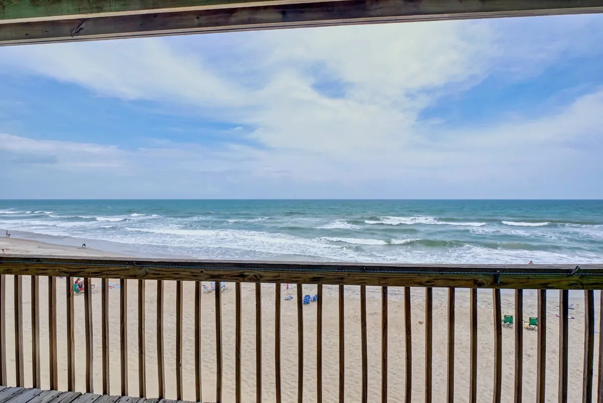 The image showcases a tranquil panorama from the condo's private deck, with stunning views of rolling Atlantic waves meeting the sandy shoreline. The wooden deck railing frames an inviting scene of endless blue sky and sparkling ocean, evoking a sense of peaceful escape and relaxation. The vantage point suggests the perfect spot for watching sunsets, beachcombing, and soaking in the coastal ambiance.