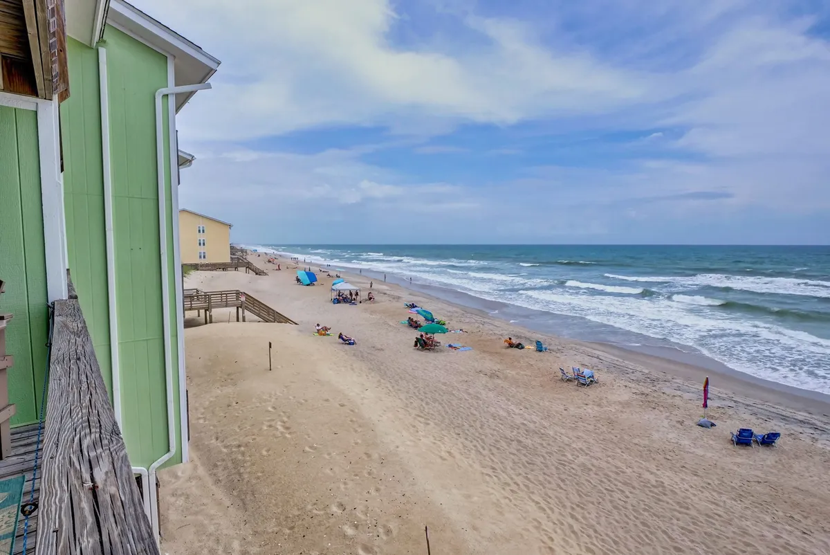 A scenic exterior view of Surf City's sandy beach with gentle waves rolling in. People are scattered along the shore, sunbathing, swimming, and playing beach games. A green building can be seen in the background, contributing to the lively, social atmosphere.