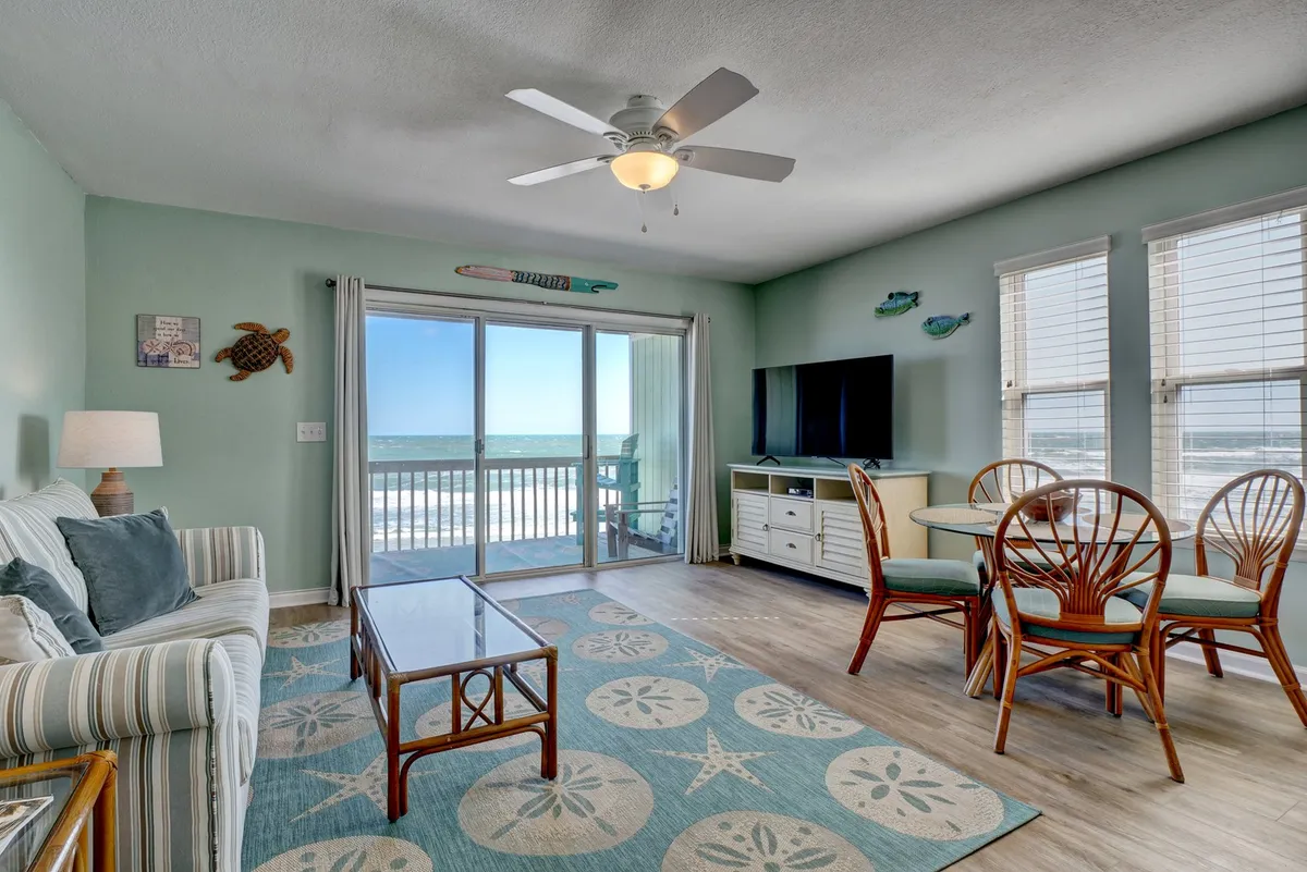 A spacious living area with a large sliding glass door that opens onto a balcony offering expansive ocean views. The room includes a comfortable striped sofa, a dining table with rattan chairs, and beach-themed wall art, creating a relaxed, light-filled environment that connects seamlessly with the outdoors.