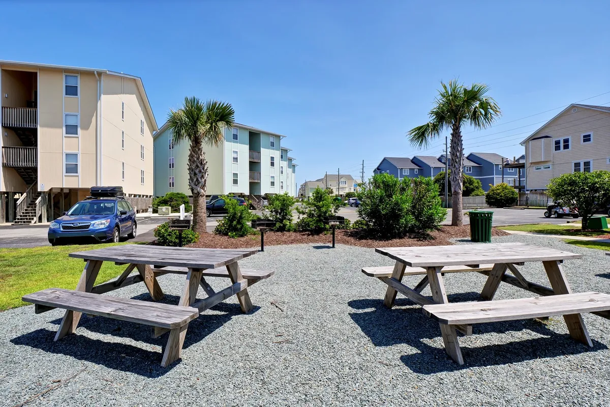 An outdoor communal space featuring picnic tables shaded by tall palm trees, adjacent to the residential buildings. The area is set up for group barbecues, casual outdoor dining, and socializing among guests.