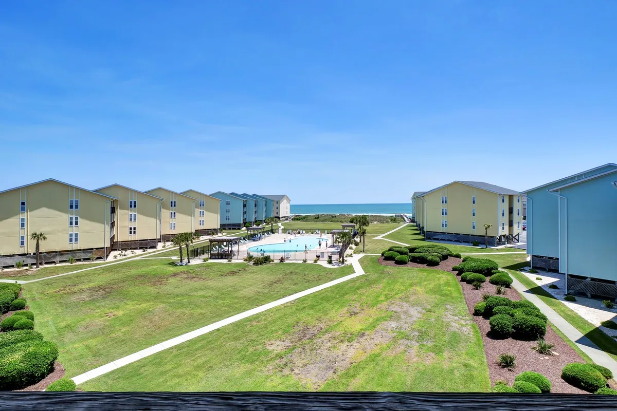 An inviting view of the community's zero-entry swimming pool, surrounded by green lawns and with a glimpse of the beach and nearby buildings in the background. The pool area is ideal for both sunbathing and socializing after a day at the beach.