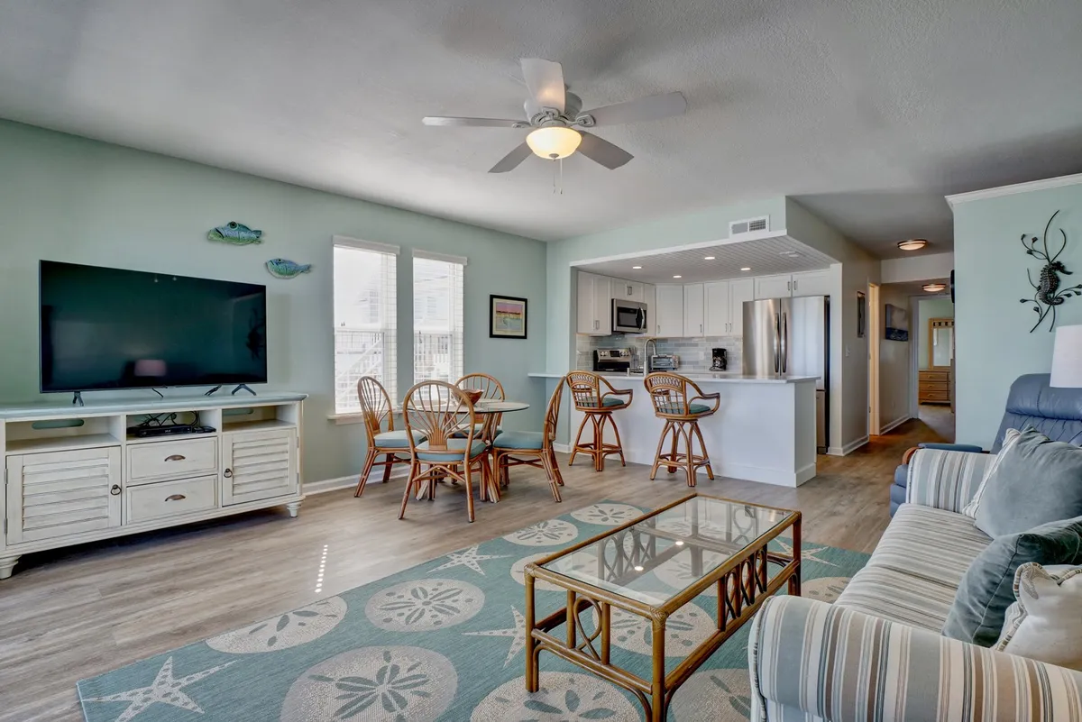 A wide-angle view of the open-plan living, dining, and kitchen area. The image features a comfortable sofa and coffee table in the foreground, a dining table for four set neatly nearby, and the kitchen area visible in the background, complete with modern appliances. Sunlight streams in, highlighting the inviting and spacious nature of the main gathering hub.